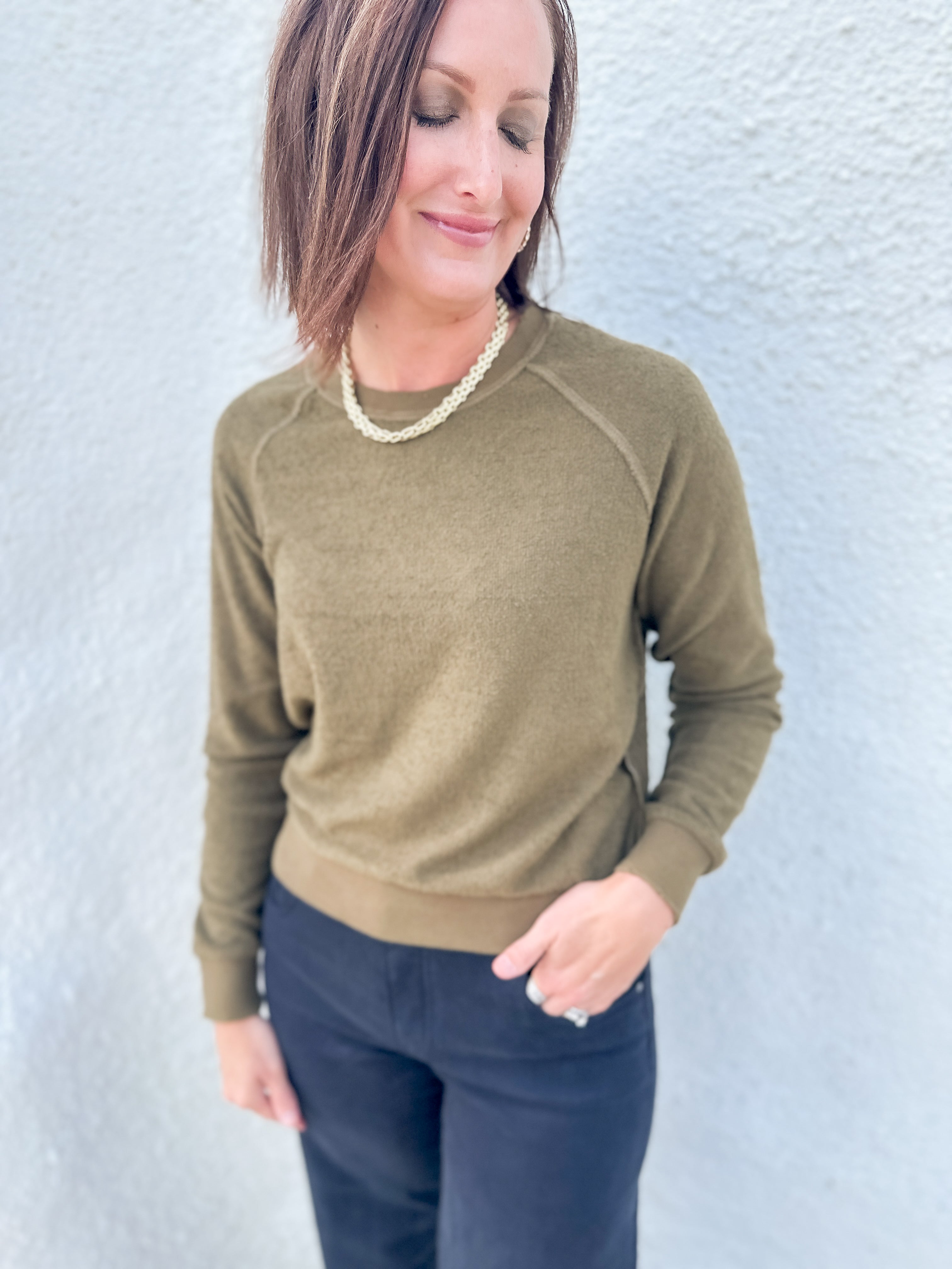A woman wearing an olive green long-sleeve crewneck sweater and dark navy pants, standing against a light gray textured wall.