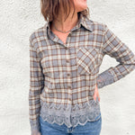A woman with a plaid flannel with lace cuff and hem details wearing blue denim pants against a white wall.