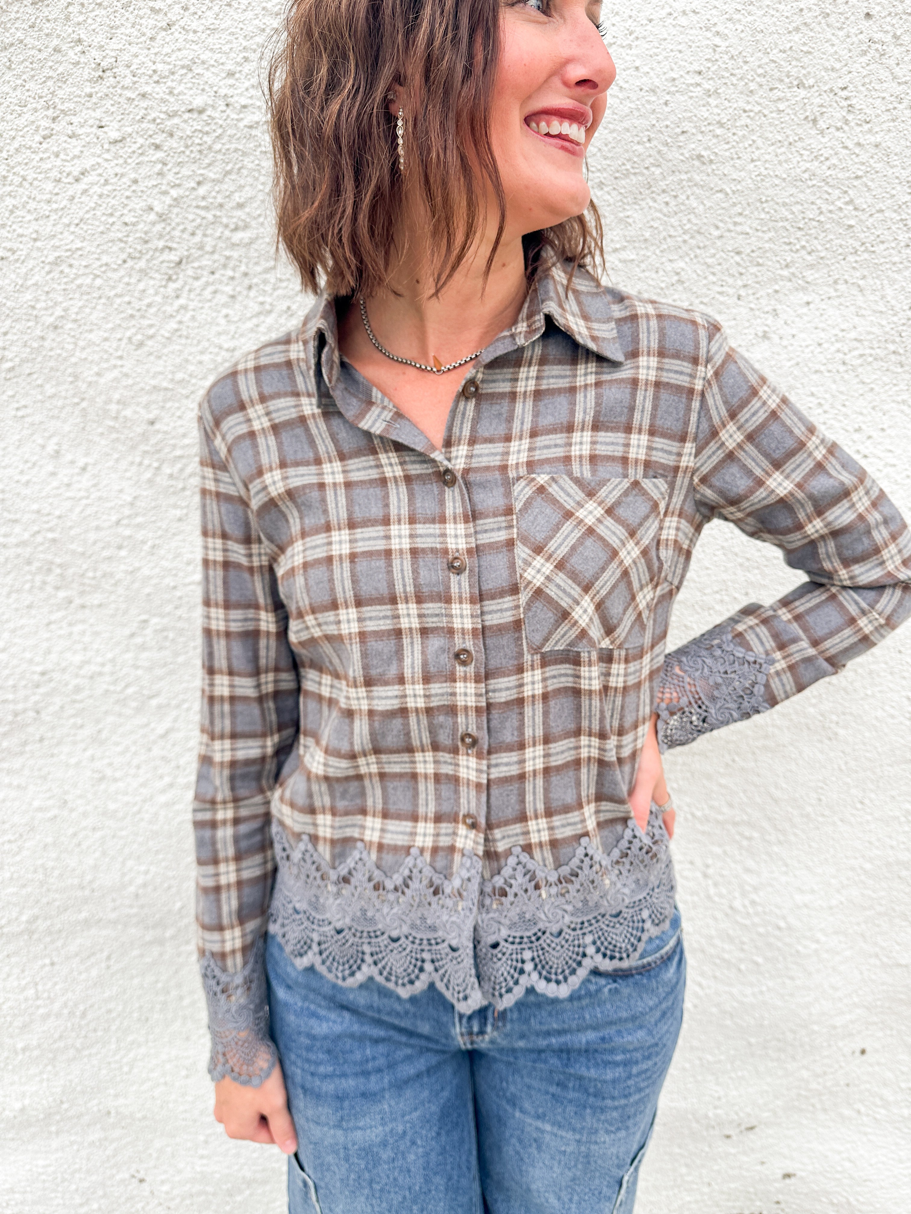 A woman with a plaid flannel with lace cuff and hem details wearing blue denim pants against a white wall.