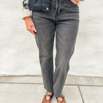 A person wearing dark gray high-waisted straight-leg jeans, a navy blue leather vest, a cream ribbed sweater, and brown leather Mary Jane flats, standing against a white stucco wall.