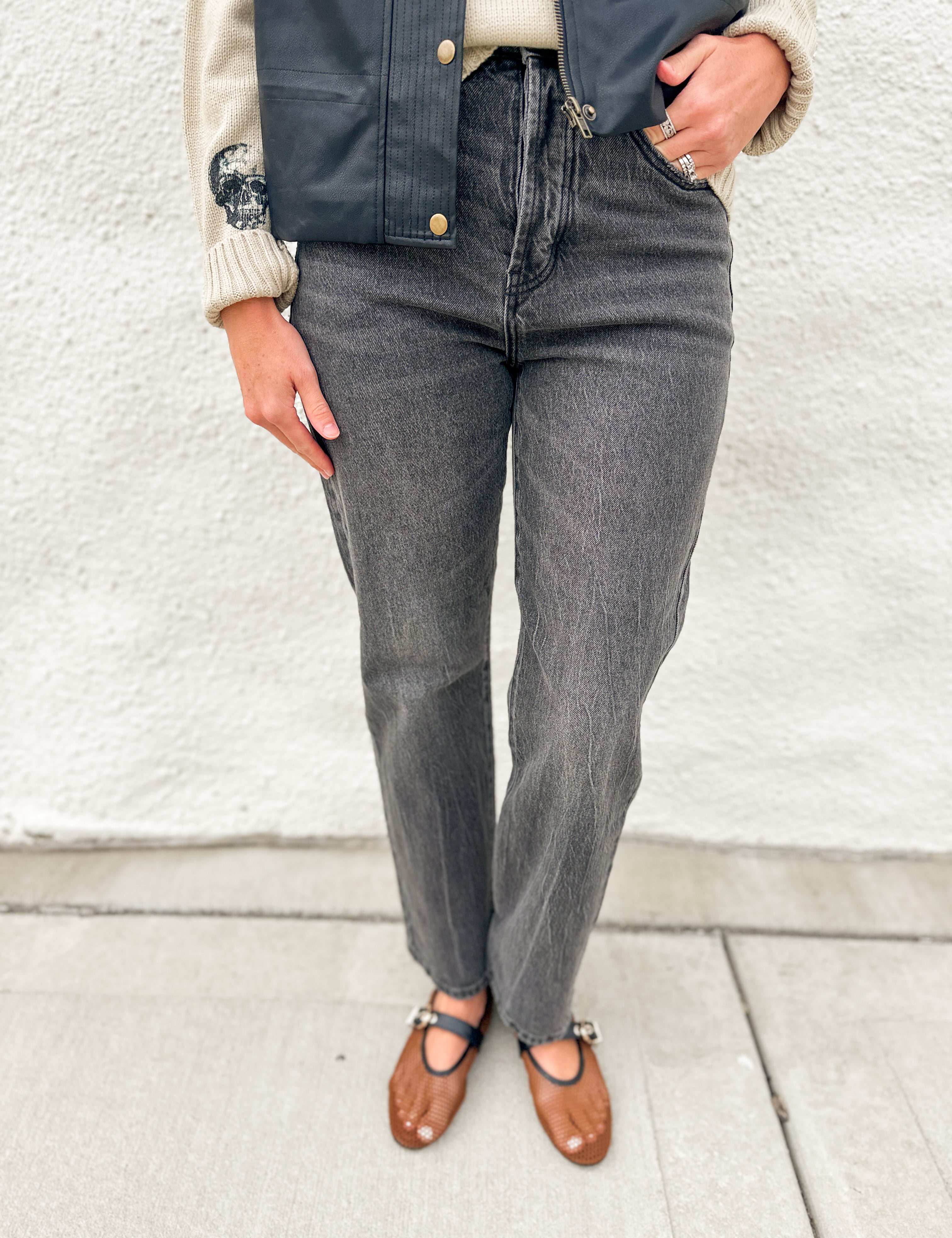 A person wearing dark gray high-waisted straight-leg jeans, a navy blue leather vest, a cream ribbed sweater, and brown leather Mary Jane flats, standing against a white stucco wall.
