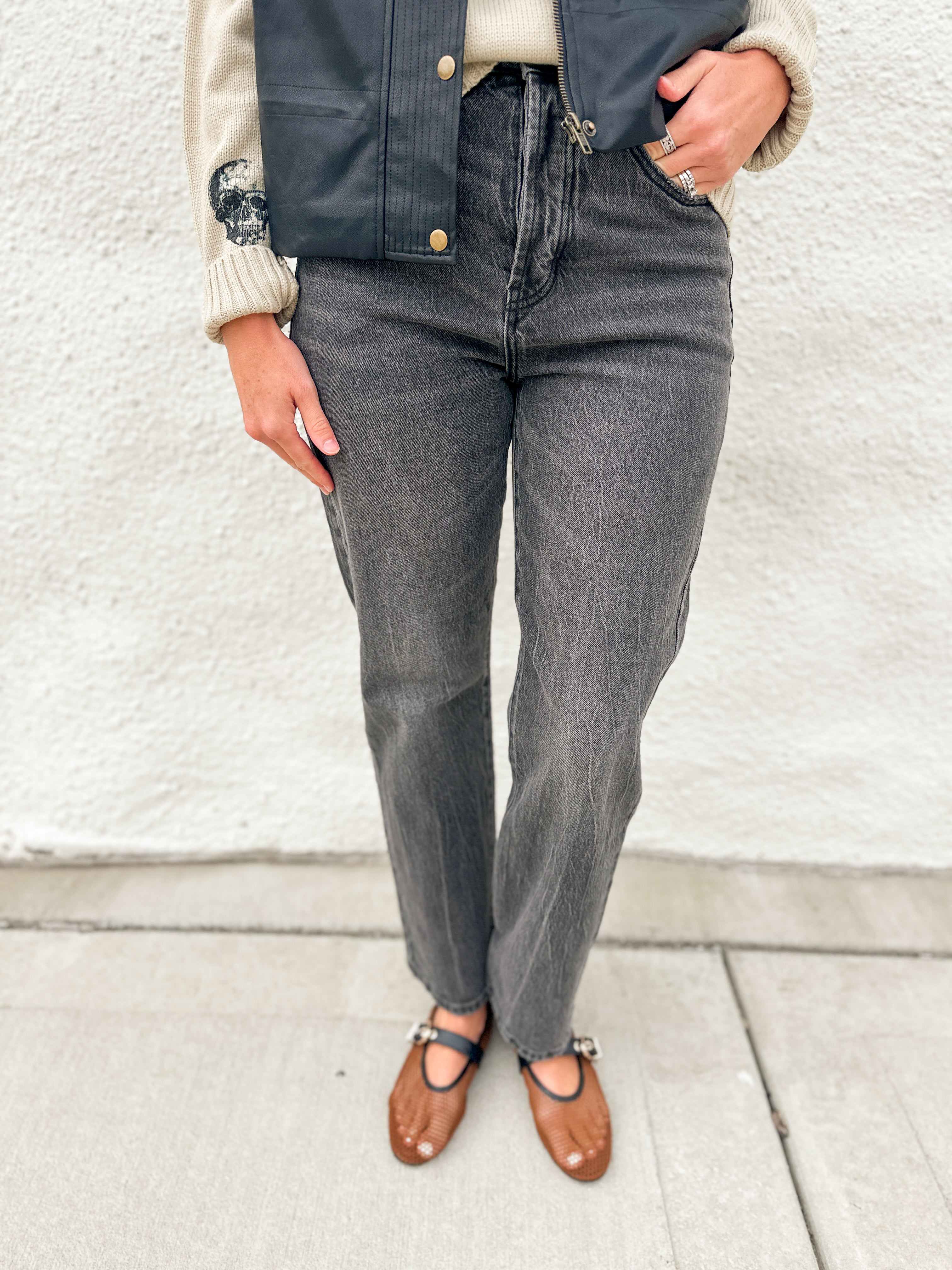A person wearing dark gray high-waisted straight-leg jeans, a navy blue leather vest, a cream ribbed sweater, and brown leather Mary Jane flats, standing against a white stucco wall.