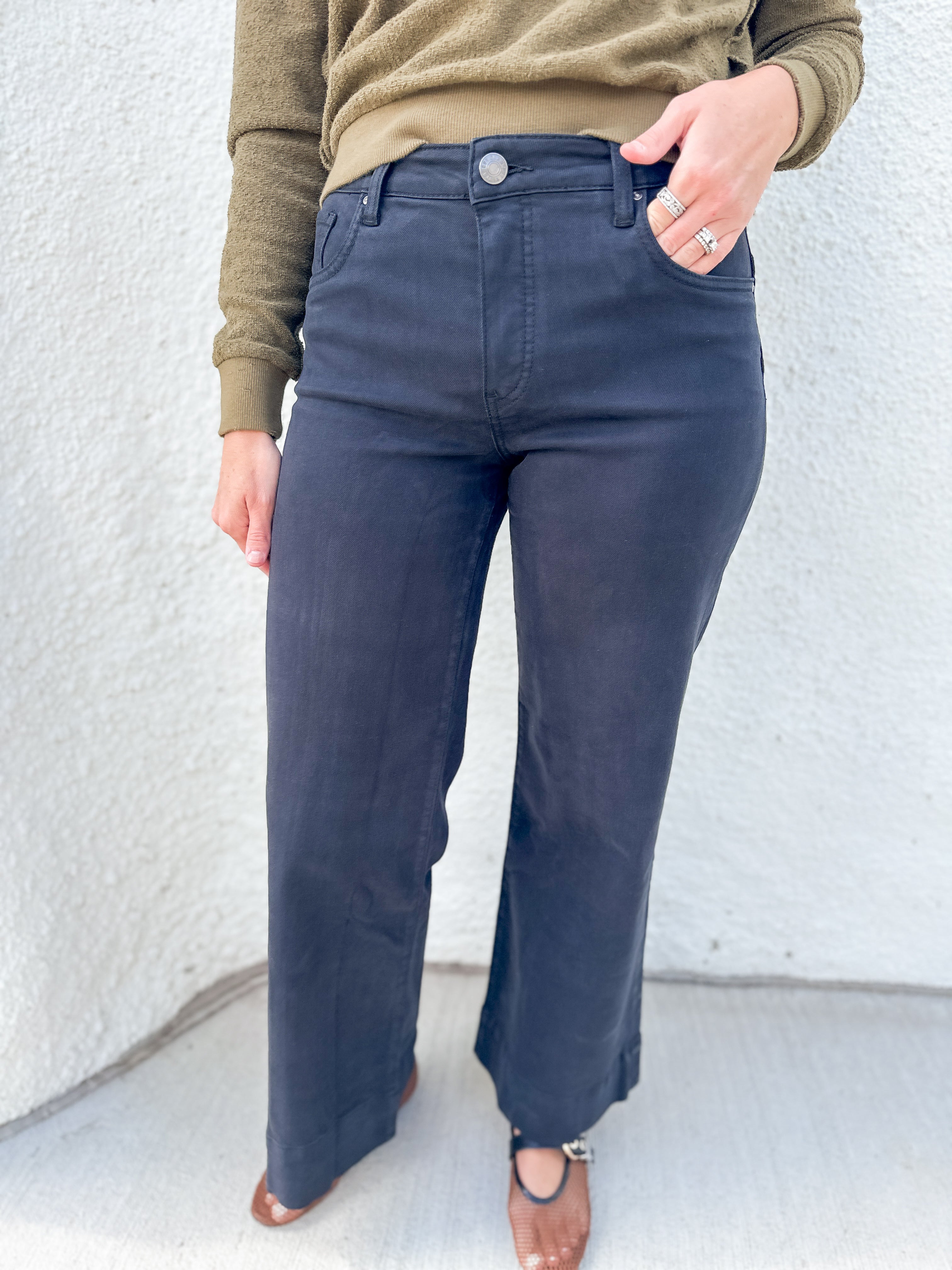A person wearing black wide-leg jeans, an olive green long-sleeve top, and brown mesh flats in a Mary Jane shoe style, standing against a white textured wall.