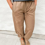 Woman wearing an embroidered faux leather crewneck tee shirt with adjustable side cinch joggers with elastic waistband and drawstring tie in a camel color and faux snakeskin clogs with gold stud detail