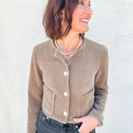 A woman wearing a cropped, waffle-knit taupe cardigan with cream buttons and front pockets, paired with dark blue jeans, standing against a textured white wall.