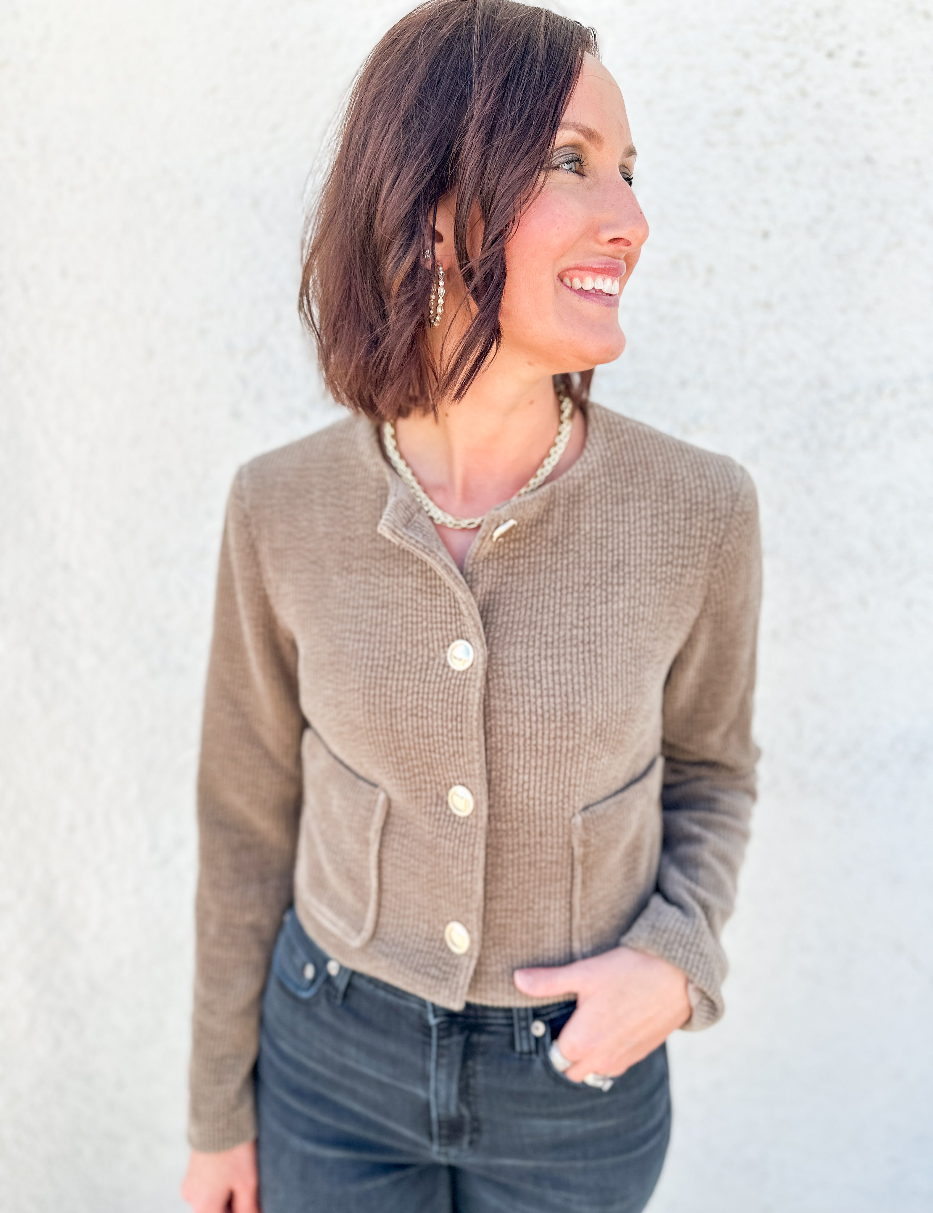 A woman wearing a cropped, waffle-knit taupe cardigan with cream buttons and front pockets, paired with dark blue jeans, standing against a textured white wall.