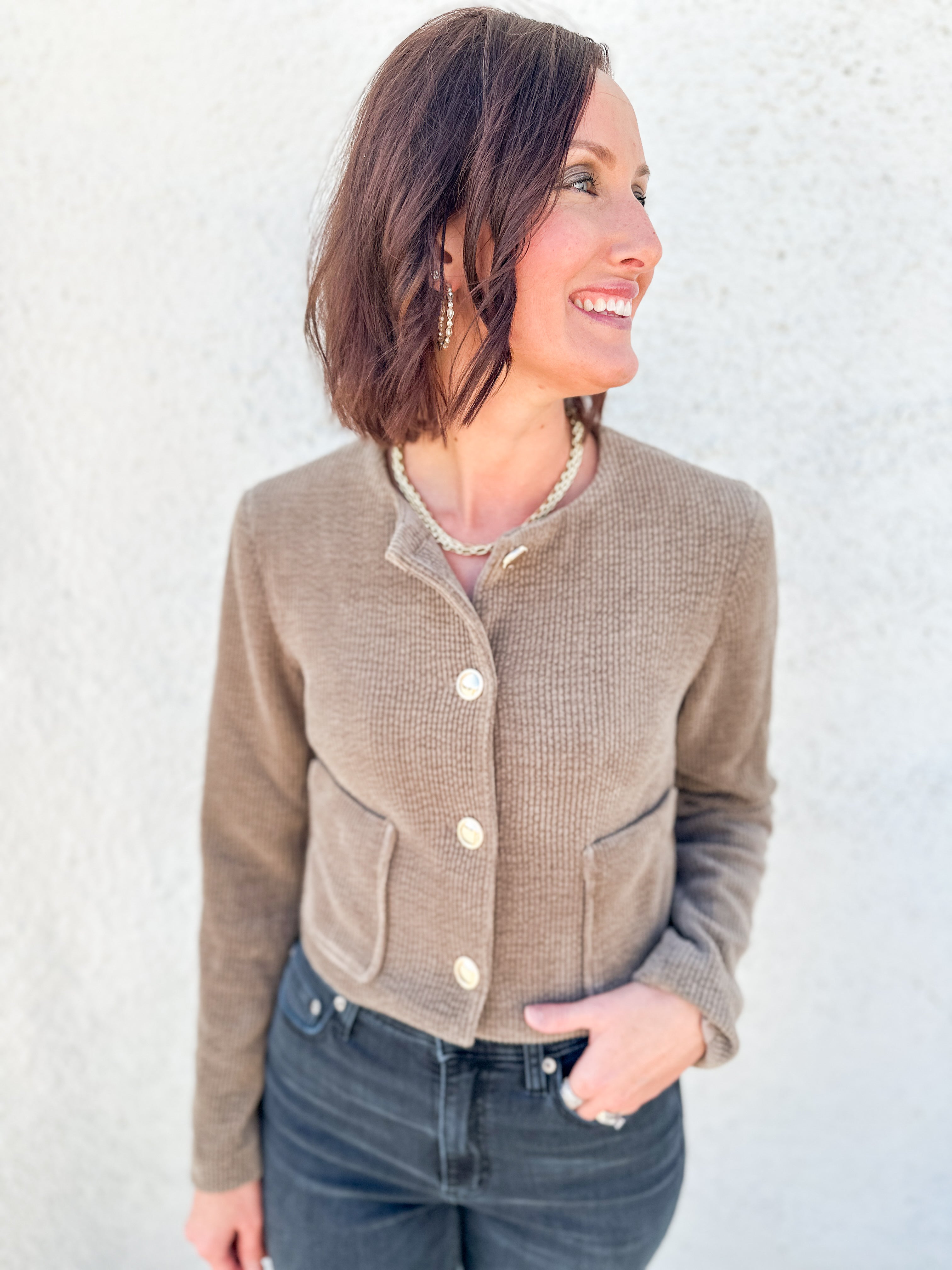 A woman wearing a cropped, waffle-knit taupe cardigan with cream buttons and front pockets, paired with dark blue jeans, standing against a textured white wall.