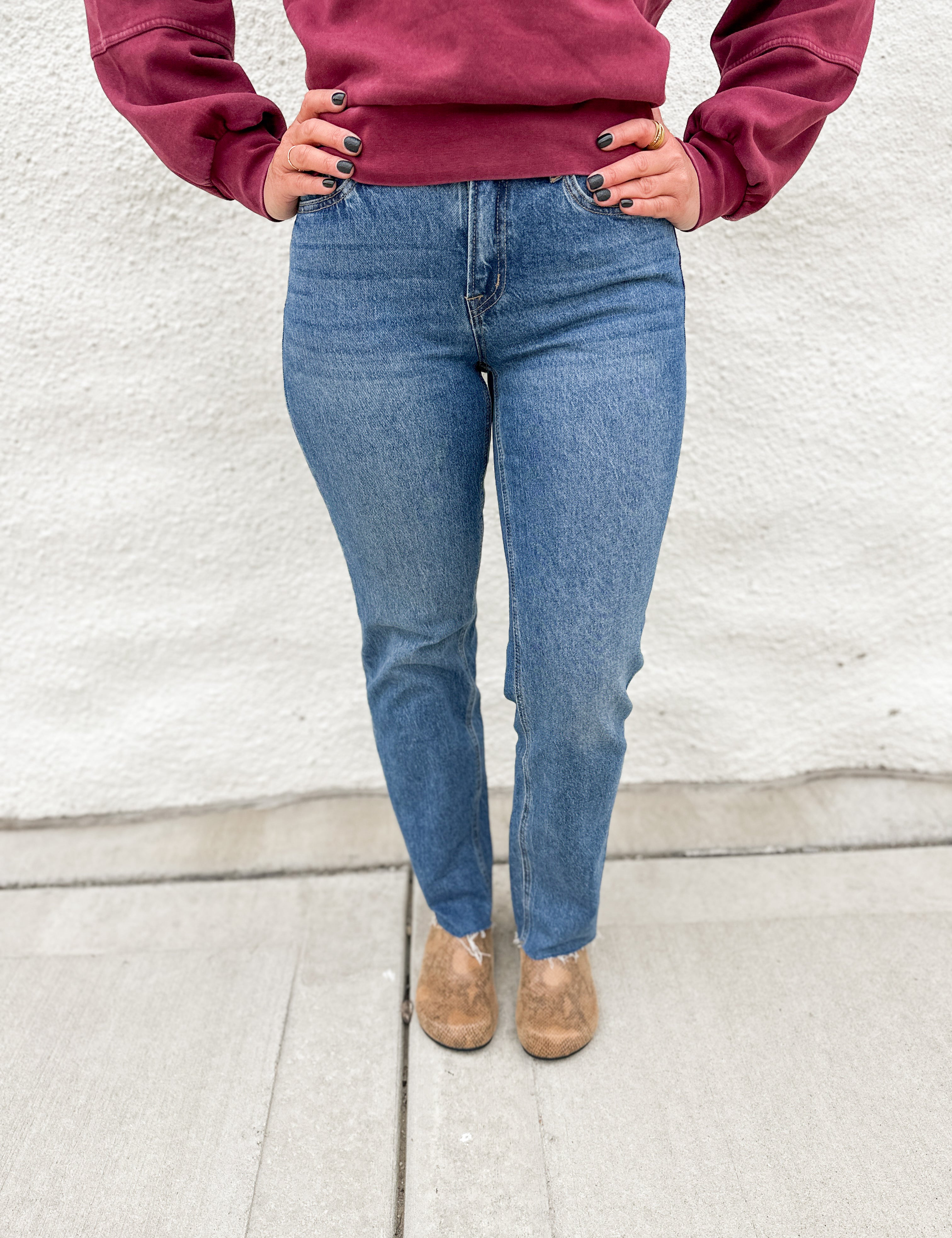 Woman wearing straight leg denim with a raw hem in a classic medium stonewash with a crewneck sweatshirt in a washed cranberry color and faux snakeskin clogs with gold studs