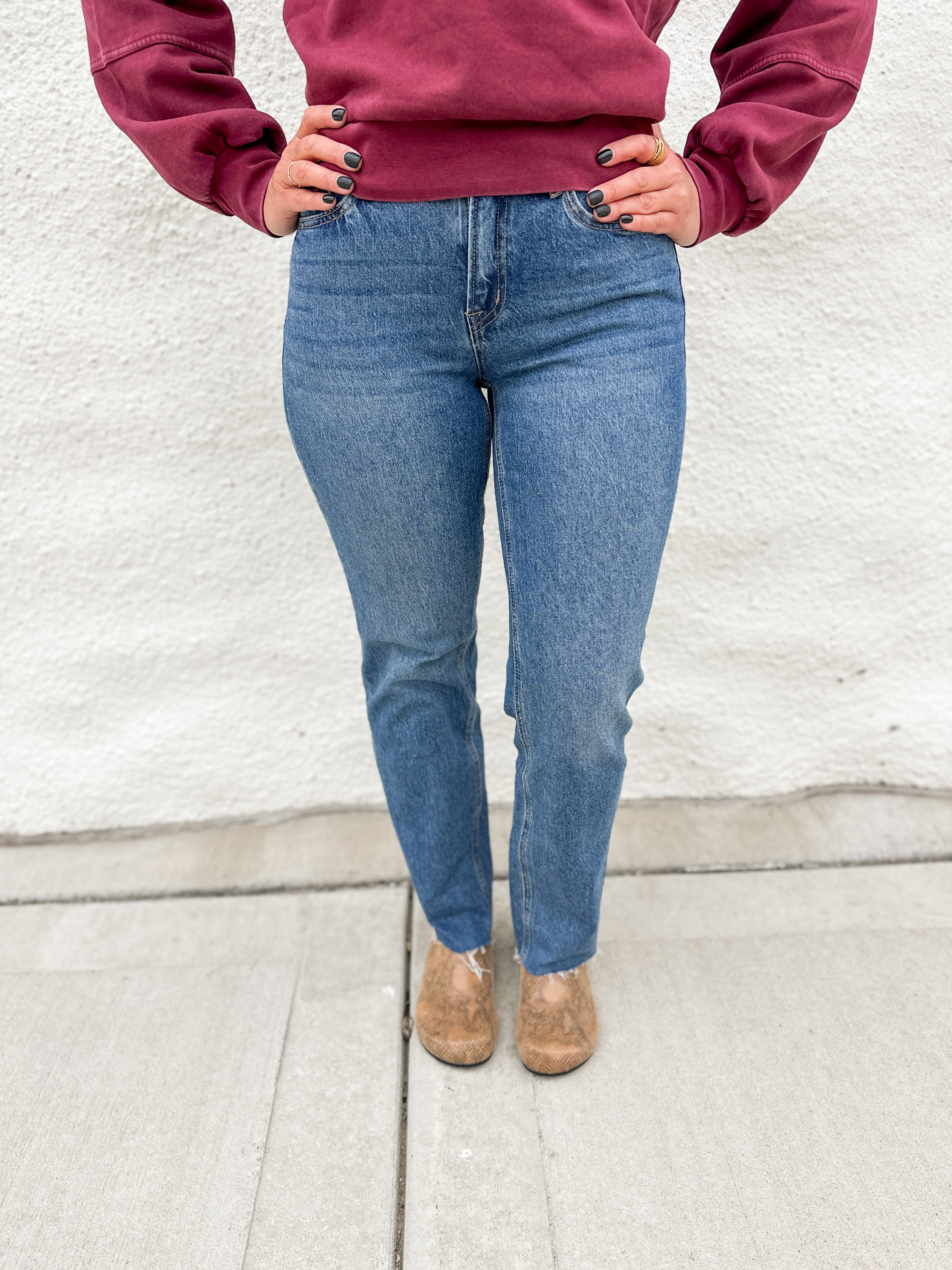 Woman wearing straight leg denim with a raw hem in a classic medium stonewash with a crewneck sweatshirt in a washed cranberry color and faux snakeskin clogs with gold studs
